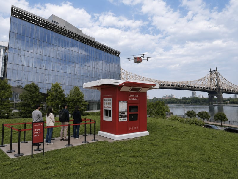 Cornell Tech receiving station on the Roosevelt Island lawn, with a raised Cornell-red landing platform, pickup lockers, and an organized waiting area separated from the drone pad.